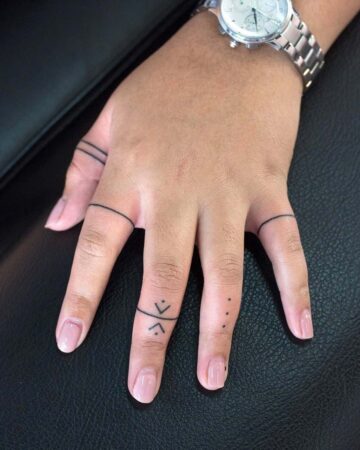 Close-up of a hand with delicate finger tattoos. The tattoos include thin bands and simple geometric designs, showcasing minimalist tattoo trends. A silver watch is visible on the wrist. The hand rests on a dark surface. Done by tattoo artists in our fine line tattoo studio parlour shop Canggu, Bali