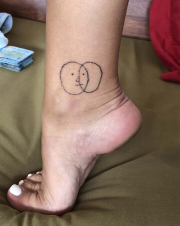 A person's foot and ankle with a minimalist ankle tattoo design of two overlapping circles, one with a simple face. The foot is flexed, showing the arch and white toenails. The background is a green cloth. This simple ankle tattoo is a great idea. Done by tattoo artists in our fine line tattoo studio parlour shop Canggu, Bali
