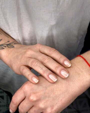 Close-up of a woman's hand with a delicate constellation tattoo on her finger, soft nail polish, and a red string bracelet on her wrist. A subtle floral tattoo is visible on her forearm.