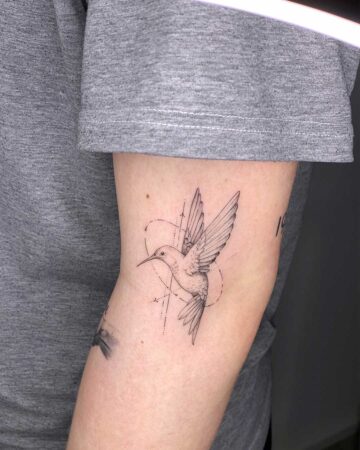 Delicate hummingbird tattoo on an arm, featuring fine line details and geometric shapes. The bird is depicted mid-flight with wings spread, set against a light gray t-shirt sleeve.