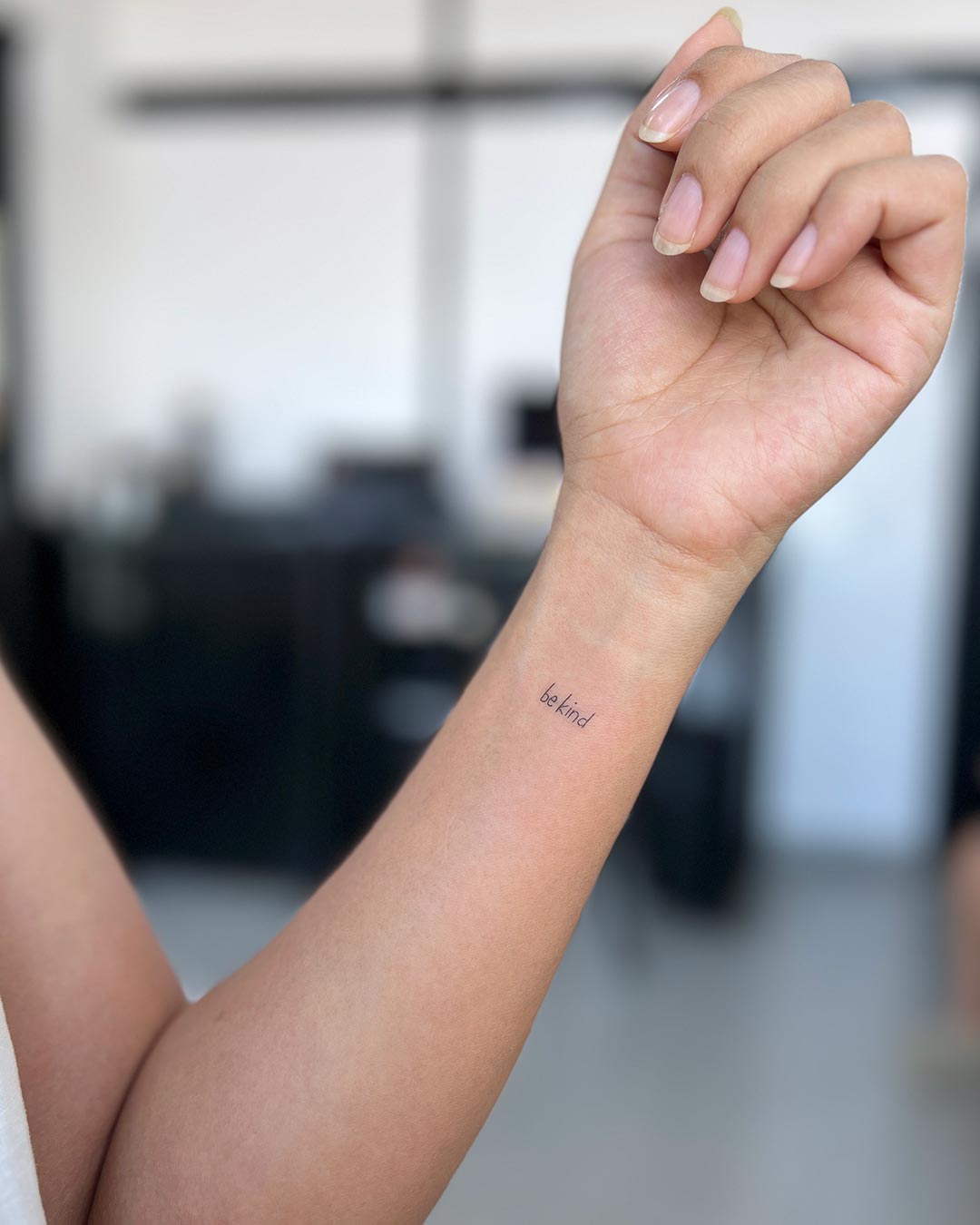 Close-up of a person's wrist with a small "be kind" tattoo in delicate script. The hand is relaxed, with neatly manicured nails, set against a blurred, bright background, evoking a sense of peace and well-being. Done by tattoo artists in our fine line tattoo studio parlour shop Canggu, Bali