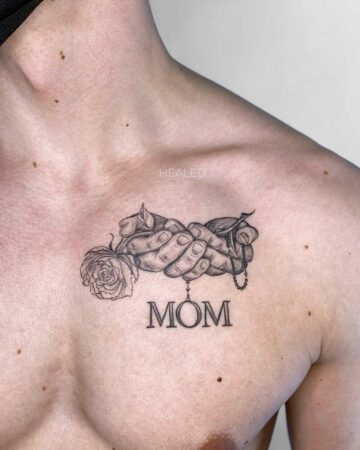 A detailed chest tattoo in black ink features clasped hands holding a rose and a rosary, with the word "MOM" beneath. A loving memorial tattoo idea for a mother. Done by tattoo artists in our fine line tattoo studio parlour shop Canggu, Bali
