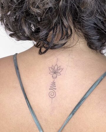 Delicate lotus flower tattoo with unalome symbol on a woman's back. Minimalist tattoo design, a popular back tattoo. The woman has short, curly hair and a gray top. Done by tattoo artists in our fine line tattoo studio parlour shop Canggu, Bali