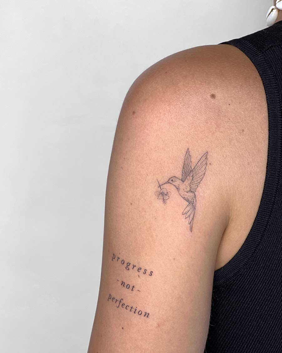 Delicate hummingbird tattoo on a woman's upper arm, holding a small flower. The tattoo is accompanied by the words "progress - not - perfection" in a simple font, embodying inspirational tattoo ideas. Done by tattoo artists in our fine line tattoo studio parlour shop Canggu, Bali