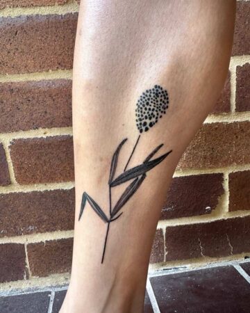 Minimalist tattoo of a bottlebrush flower on a leg, featuring dotted texture on the flower head and linear leaf details. Brick wall background.