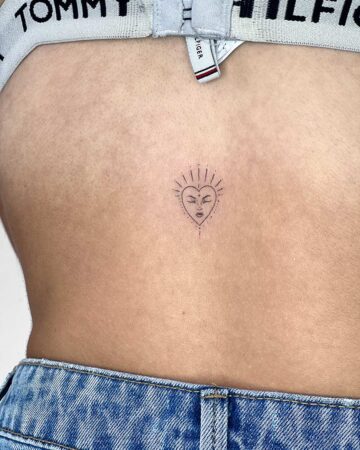 Delicate minimalist heart tattoo on a woman's back. The design features a heart shape with sun rays, a face, and fine line details, a perfect small tattoo idea. Done by tattoo artists in our fine line tattoo studio parlour shop Canggu, Bali
