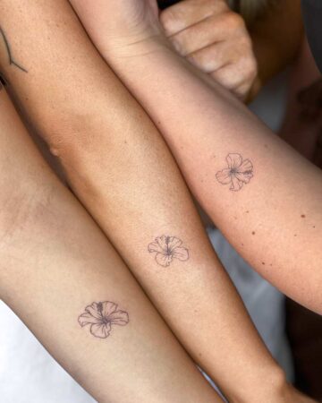 Three arms with matching hibiscus flower tattoos, a symbol of friendship. The delicate, fine-line designs adorn the forearms, representing a strong bond between friends. The tattoos are a perfect example of friendship tattoos and flower tattoos. Done by tattoo artists in our fine line tattoo studio parlour shop Canggu, Bali