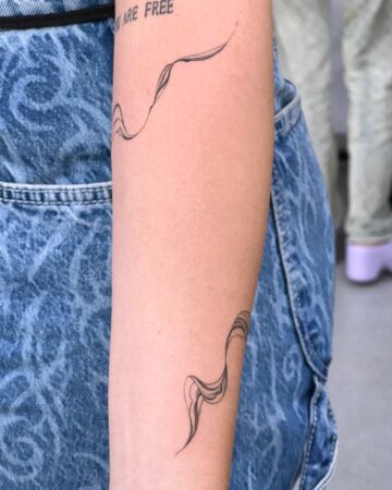 Delicate fine line arm tattoo featuring two flowing, ribbon-like designs. The tattoo is inked in black on fair skin, with the words "ARE FREE" tattooed above. A minimalist tattoo idea. Done by tattoo artists in our fine line tattoo studio parlour shop Canggu, Bali