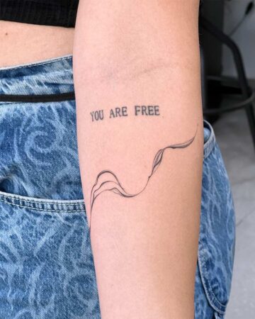 A delicate, fine-line minimalist tattoo on an arm reads "YOU ARE FREE" above a flowing, abstract design. The tattoo is on fair skin, with blue jeans visible in the background, showcasing a small tattoo design. Done by tattoo artists in our fine line tattoo studio parlour shop Canggu, Bali