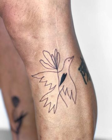 A minimalist line art dove tattoo on a leg. The design features a dove in flight with delicate wing details and a black heart shape. The small tattoo is a stylish example of minimalist tattoo art. Done by tattoo artists in our fine line tattoo studio parlour shop Canggu, Bali