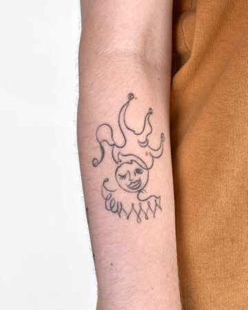 A whimsical, minimalist tattoo of a smiling jester, rendered in a single black line on a person's forearm. The design includes a winking face, a ruffled collar, and a playful crown, showcasing a unique minimalist tattoo style. Done by tattoo artists in our fine line tattoo studio parlour shop Canggu, Bali