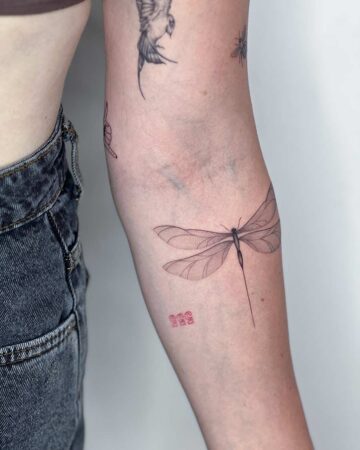 Delicate dragonfly tattoo on forearm, featuring fine line detail and translucent wings. A minimalist tattoo design with a long, thin abdomen. Small red "555" tattoo below, along with other small tattoos. Done by tattoo artists in our fine line tattoo studio parlour shop Canggu, Bali