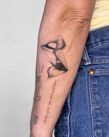 Close-up of a detailed lip tattoo on an arm, featuring two sets of lips in a kissing pose. The tattoo includes the words "Write your own masterpiece" and a small drawing of a figure smoking a cigarette. A stylish tattoo design, perfect for tattoo inspiration. Done by tattoo artists in our fine line tattoo studio parlour shop Canggu, Bali