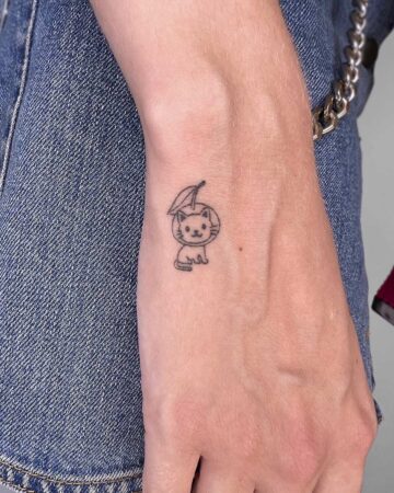 A tiny, minimalist cat tattoo on a person's wrist. The black outline depicts a cat wearing a cherry on its head, sitting upright. The tattoo is simple and delicate, a perfect small tattoo idea. Done by tattoo artists in our fine line tattoo studio parlour shop Canggu, Bali