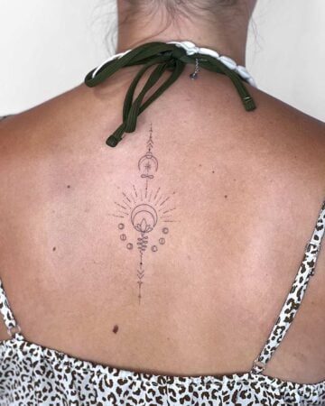 Elegant minimalist tattoo design on a woman's back. The delicate, fine-line tattoo features a crescent moon, lotus flower, sunburst, and spiritual symbols, perfectly placed. A beautiful example of back tattoo art. Done by tattoo artists in our fine line tattoo studio parlour shop Canggu, Bali