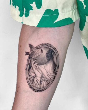 Detailed black and grey tattoo of a woman in an ornate oval frame, with a small bird perched on her hand. The artistic tattoo design is on the arm, with a green patterned shirt in the background. Done by tattoo artists in our fine line tattoo studio parlour shop Canggu, Bali