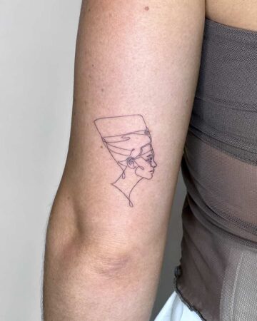 Delicate minimalist line art tattoo of Nefertiti on a woman's upper arm. The tattoo showcases a single-line design, capturing the profile of the Egyptian queen with her iconic headdress. This elegant arm tattoo is a beautiful example of minimalist tattoo art. Done by tattoo artists in our fine line tattoo studio parlour shop Canggu, Bali