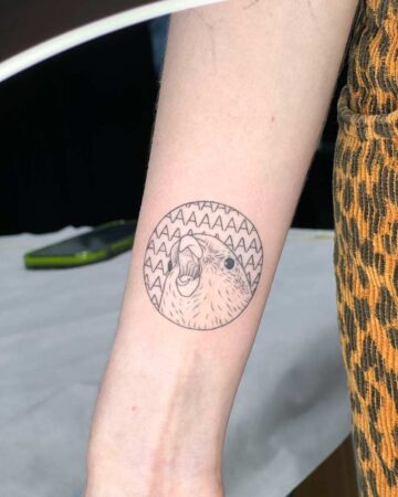 A minimalist bird tattoo on a forearm, featuring a screaming bird inside a circle with repeating "AAAAA" text. The tattoo is a fine-line design, showcasing a unique and creative tattoo idea. The arm is against a background of a leopard print fabric. Done by tattoo artists in our fine line tattoo studio parlour shop Canggu, Bali