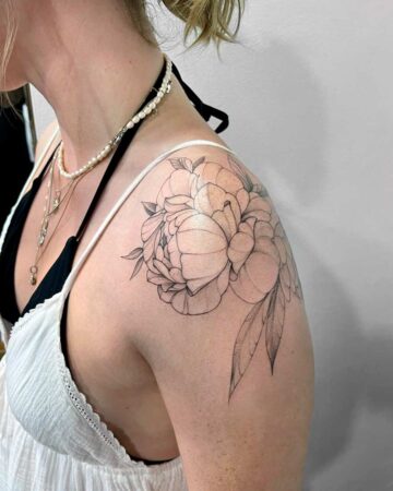 A woman's shoulder featuring a delicate peony tattoo with intricate line work. The black ink tattoo showcases the flower's petals and leaves in a detailed design, a beautiful example of a shoulder tattoo. Done by tattoo artists in our fine line tattoo studio parlour shop Canggu, Bali