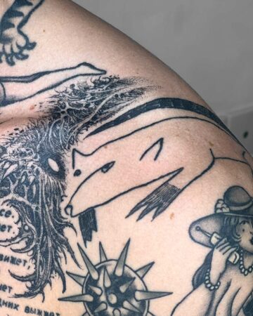 Close-up of a person's shoulder covered in various black ink tattoos. A stylized wolf or fox is prominent, along with other designs including a spiky shape and a woman. The body art showcases a range of styles and themes. Done by tattoo artists in our fine line tattoo studio parlour shop Canggu, Bali
