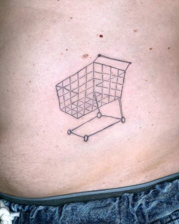A minimalist shopping cart tattoo, rendered in fine black lines, sits on a person's torso just above the waistband of their jeans. The tattoo design features the cart's wireframe structure, showcasing a unique body art choice. Done by tattoo artists in our fine line tattoo studio parlour shop Canggu, Bali