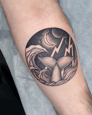 A detailed black ink tattoo on an arm depicts a whale's tail emerging from stormy waves within a circle. Lightning flashes against a night sky with a crescent moon, a striking example of tattoo designs. Done by tattoo artists in our fine line tattoo studio parlour shop Canggu, Bali