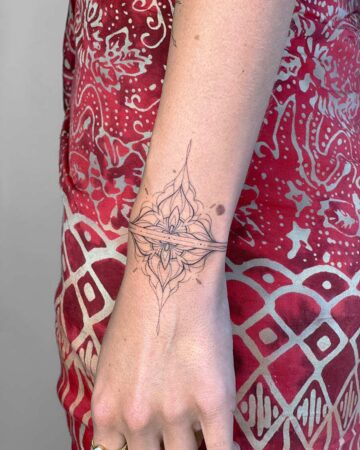 A close-up of a delicate floral tattoo on a wrist. The fine line tattoo features a symmetrical design with a central flower and surrounding leaves, appearing like a bracelet. The person wears a red patterned top. Done by tattoo artists in our fine line tattoo studio parlour shop Canggu, Bali