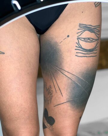 Close-up of a person's thigh featuring a bold, shaded black tattoo with geometric lines and a minimalist eye design. Other small tattoos are visible. The skin contrasts with the dark ink, showcasing modern body art. Done by tattoo artists in our fine line tattoo studio parlour shop Canggu, Bali