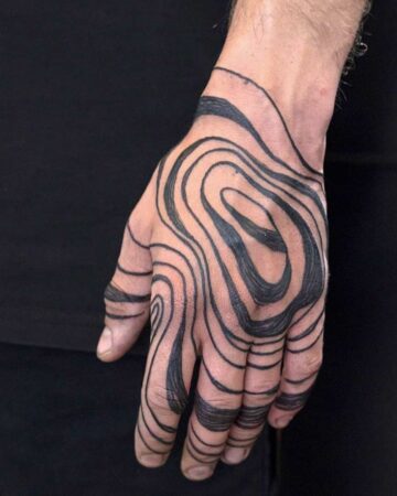 Close-up of a hand covered in a black, abstract tattoo design. The swirling lines create a topographic map effect, covering the palm and fingers. A striking example of hand tattoo body art. Done by tattoo artists in our fine line tattoo studio parlour shop Canggu, Bali