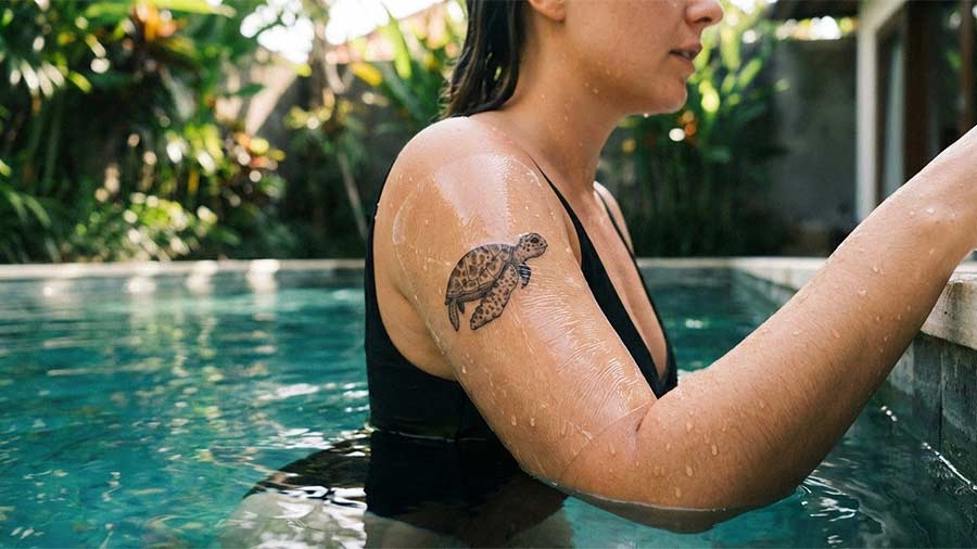 Woman with a turtle tattoo covered with second skin on her arm, relaxing in a pool. This style reflects the artistry beyond fine line tattoos. Created by tattoo artists in Canggu, Bali. Bali tattoo design.