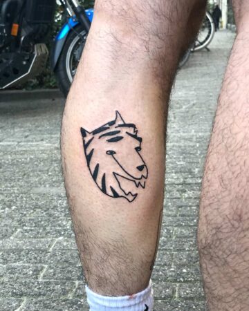 A black ink tiger tattoo on a hairy leg. The cartoonish tiger head has striped markings and a smiling expression, a fun piece of tattoo art. Done by tattoo artists in our fine line tattoo studio parlour shop Canggu, Bali