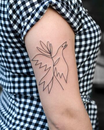 A minimalist line art bird tattoo on a woman's upper arm. The black ink design features a bird with outstretched wings and simple, stylized leaves, set against a checkered shirt, showcasing a stylish minimalist tattoo idea.