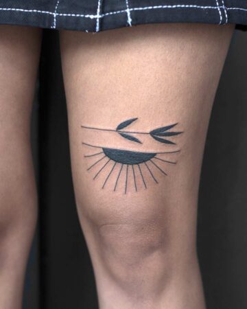 A minimalist tattoo design of an eye with sun rays and a branch on a woman's thigh. The black ink is a simple, elegant design for a small tattoo idea. Done by tattoo artists in our fine line tattoo studio parlour shop Canggu, Bali