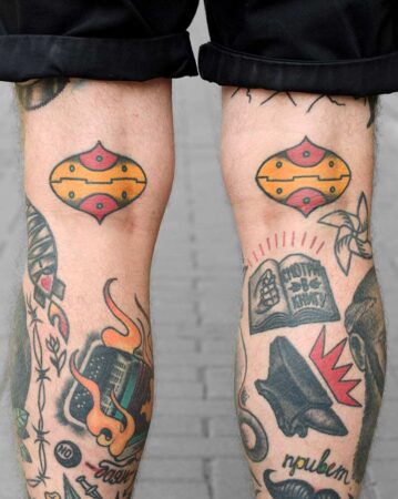 Close-up of a person's legs covered in colorful tattoos, showcasing various tattoo art designs. The knees feature matching geometric ink designs. Other tattoos include a burning accordion and a book with Russian text. The word "привет" is also visible. Done by tattoo artists in our fine line tattoo studio parlour shop Canggu, Bali