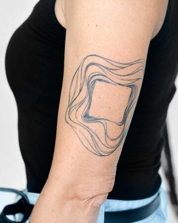 A woman's arm displays an abstract tattoo design. The tattoo features a central, slightly irregular square surrounded by flowing, wavy lines in dark blue ink, creating a minimalist aesthetic. This geometric tattoo is an example of abstract tattoo art. Done by tattoo artists in our fine line tattoo studio parlour shop Canggu, Bali