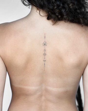 A woman's bare back with a delicate spine tattoo featuring a lotus flower, an Om symbol, and other geometric designs. The tattoo reflects a focus on yoga, meditation, and mindfulness, embracing body art. Done by tattoo artists in our fine line tattoo studio parlour shop Canggu, Bali
