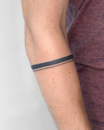 Close-up of a man's forearm with a minimalist armband tattoo. The black tattoo band is composed of two lines, a thicker one above a thinner one. The tattoo is on light skin, with a burgundy shirt in the background. Simple tattoo ideas for men. Done by tattoo artists in our fine line tattoo studio parlour shop Canggu, Bali