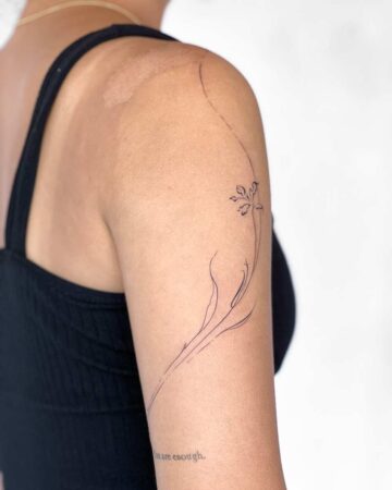 Delicate minimalist floral tattoo design on an arm, featuring a thin, curved vine with a small flower. The black ink tattoo is on a woman's arm against a plain white background. Done by tattoo artists in our fine line tattoo studio parlour shop Canggu, Bali