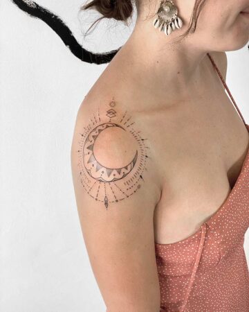 A woman with a minimalist moon tattoo on her shoulder, featuring a crescent moon, dotted lines, and a sunburst design. The tattoo is black and white, and the woman wears a peach-colored dress. This moon tattoo is a beautiful example of a celestial tattoo. Done by tattoo artists in our fine line tattoo studio parlour shop Canggu, Bali