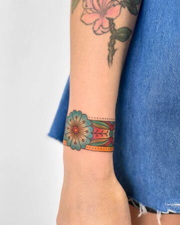 Vibrant wrist tattoo featuring a large, stylized blue flower and a colorful geometric band. The tattoo design showcases bright red, green, and yellow hues, creating a striking floral tattoo. The arm is partially visible, with a faded pink flower tattoo on the upper arm.