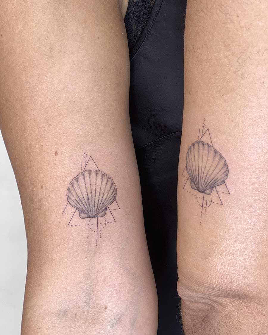 Two arms side-by-side, each displaying a matching minimalist sea shell tattoo. The delicate line art depicts a scallop shell within a geometric triangle and lines, perfect for couples tattoos. The design is a beautiful example of matching tattoos. Done by tattoo artists in our fine line tattoo studio parlour shop Canggu, Bali
