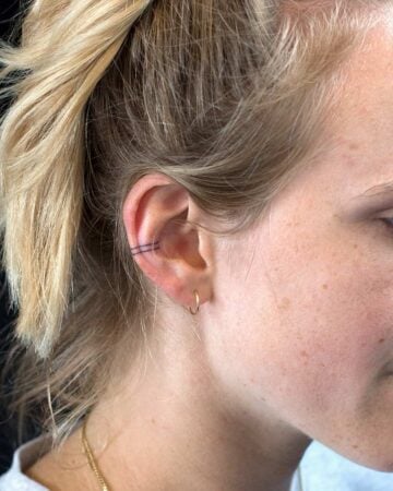 Close-up of a woman's ear with a small gold hoop earring and a minimalist ear tattoo design. Two black lines are tattooed on the helix cartilage, demonstrating a popular minimalist ear tattoo style. Done by tattoo artists in our fine line tattoo studio parlour shop Canggu, Bali
