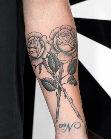 Detailed black and grey rose tattoo on a forearm, featuring two roses with intricate shading and leaves. The name "Nia" is tattooed below the roses, showcasing a classic floral design. Done by tattoo artists in our fine line tattoo studio parlour shop Canggu, Bali