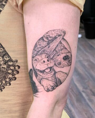 A detailed dog tattoo on an arm, showcasing a laughing dog's face, with a second dog in the background, all within a circular frame. This funny dog tattoo is a unique pet portrait. Done by tattoo artists in our fine line tattoo studio parlour shop Canggu, Bali