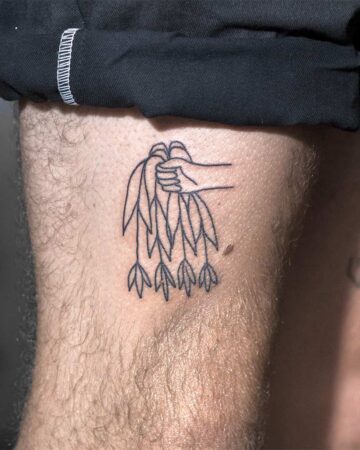 A minimalist line art tattoo of a hand holding a bundle of leaves. The tattoo is on a hairy arm, with a black shirt partially visible in the background. Simple tattoo ideas for inspiration. done by artists in our fine line tattoo studio Canguu Bali