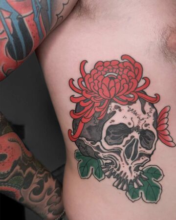 Rib tattoo of a skull with a vibrant red chrysanthemum flower on top, accented by green leaves. The skull design is detailed with shading and fine lines, part of a larger collection of body art. Japanese (Irezumi/Wabori) tattoo artist studio Canggu Bali