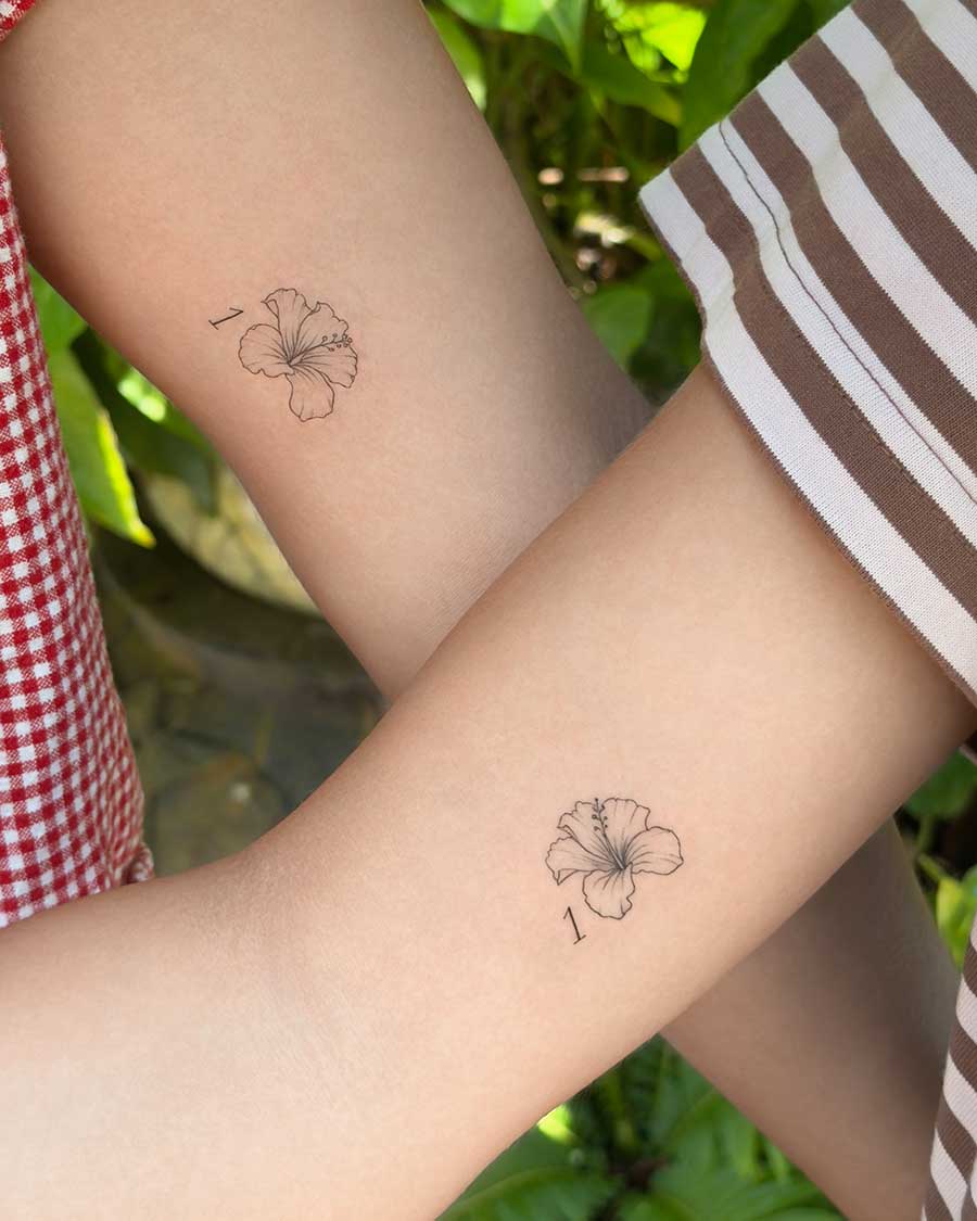 Two arms crossed to show matching minimalist hibiscus flower tattoos. The flowers are drawn with very thin lines and accompanied by a small, elegant number "1" next to each bloom.