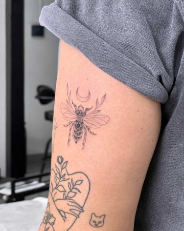 Delicate bee tattoo design on a forearm, featuring a crescent moon and floral details. The minimalist tattoo is inked in fine lines, showcasing intricate wings, and is part of a larger collection of arm tattoos. Done by tattoo artists in our fine line tattoo studio parlour shop Canggu, Bali