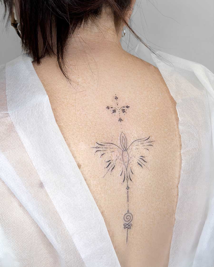 Close-up of an ornamental tattoo on a person's upper back, featuring a delicate line-art phoenix with intricate dotwork details and a geometric symbol below. This ornamental tattoo is a beautiful example of fine line artistry. Created in the multi style tattoo studio in Canggu Bali.