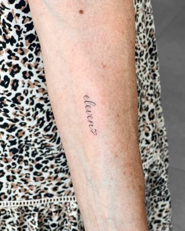 A small, delicate "eleven" script tattoo on a woman's forearm, with a heart replacing the final "n". The tattoo is centered on the arm, showcasing a minimalist design. This is a great example of a meaningful tattoo. done by artists in our fine line tattoo studio Canguu Bali